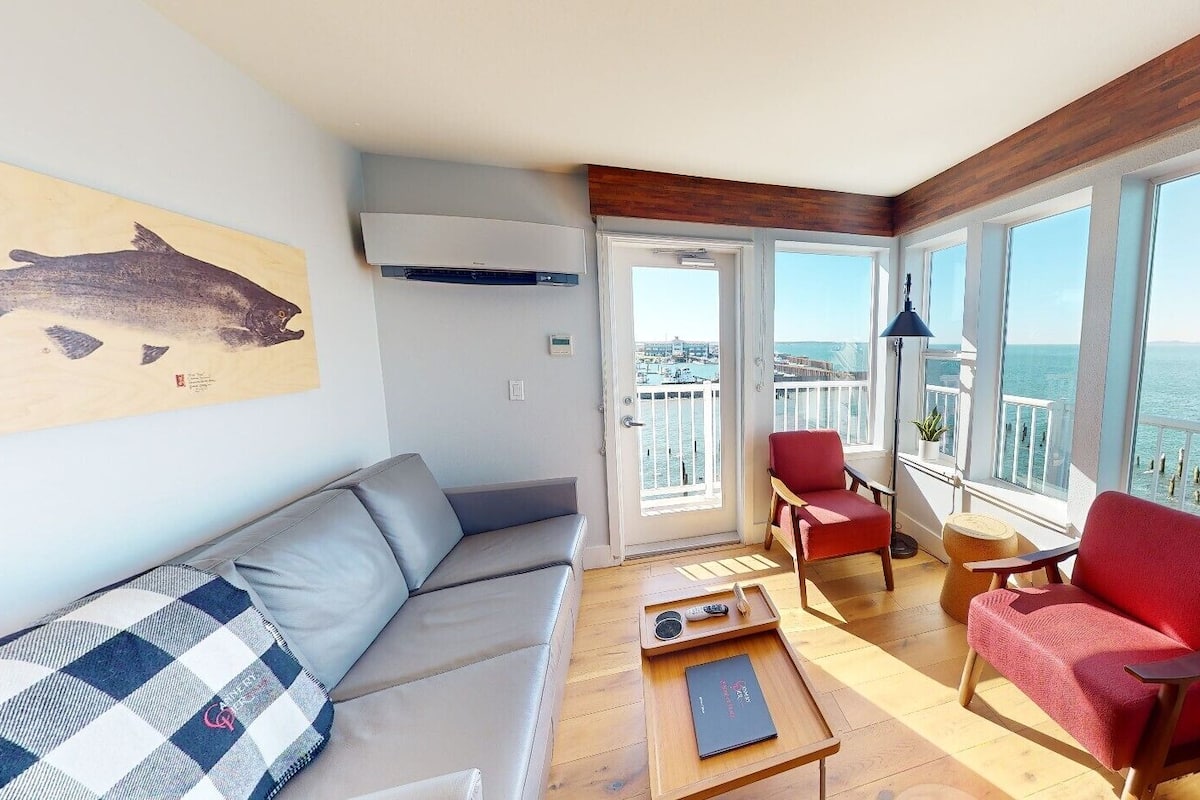 A modern living room with a sofa, red chairs, a coffee table, and a view of the sea through large windows.