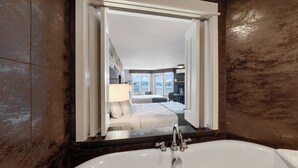 A modern bathroom with a large bathtub, a mirror reflecting a bedroom with a bed and a desk, and a view of a balcony.