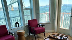 A room with large windows overlooking a bridge and water, featuring two red chairs, a small wooden stool, and a wooden table with a tray.