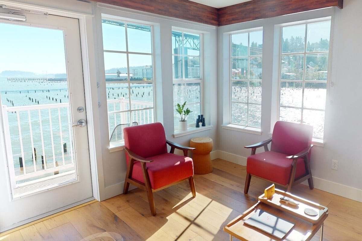 A room with large windows, two red chairs, a small table, and a view of the water.