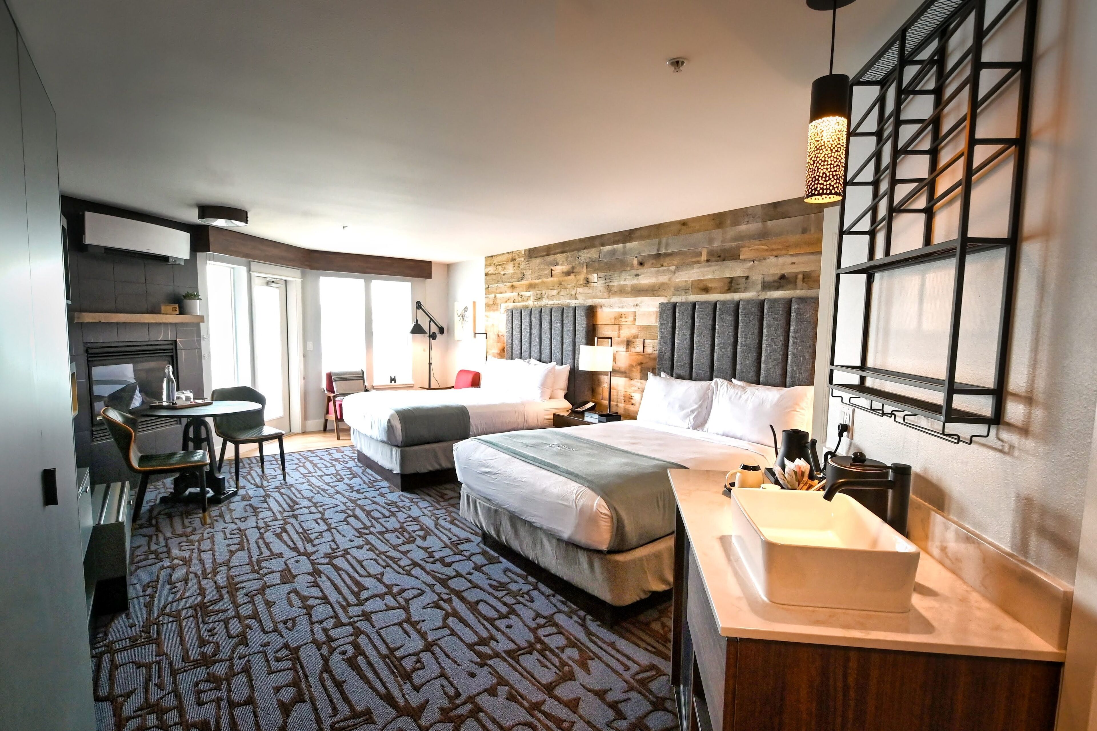 A hotel room with two beds, a desk, a chair, a large window, a wall-mounted TV, and a modern bathroom with a large sink and mirror.