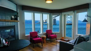 A room with a fireplace, a round table, two red chairs, a sofa, and large windows offering a view of a bridge and water.