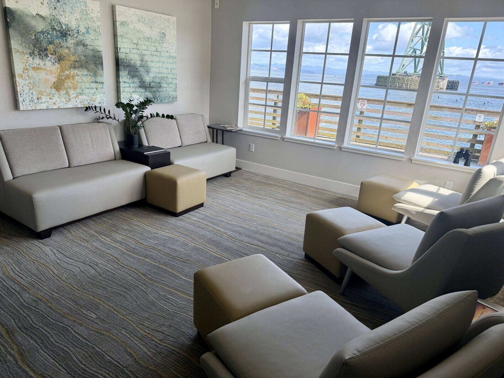 A modern living room with a sofa, ottoman, and chairs, large windows with a view of the sea and a bridge, and abstract wall art.