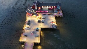 Vue aérienne d’un hôtel situé au bord de l’eau, avec un parking, un bâtiment à plusieurs étages et une jetée qui s’avance dans l’eau.