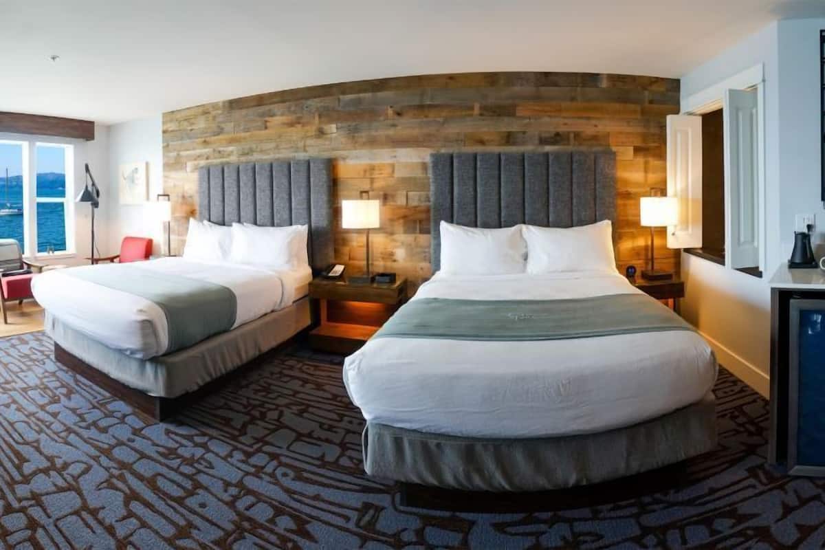 A hotel room with two beds, a curved wooden headboard, a large window with a view, a desk with a chair, and a minibar.