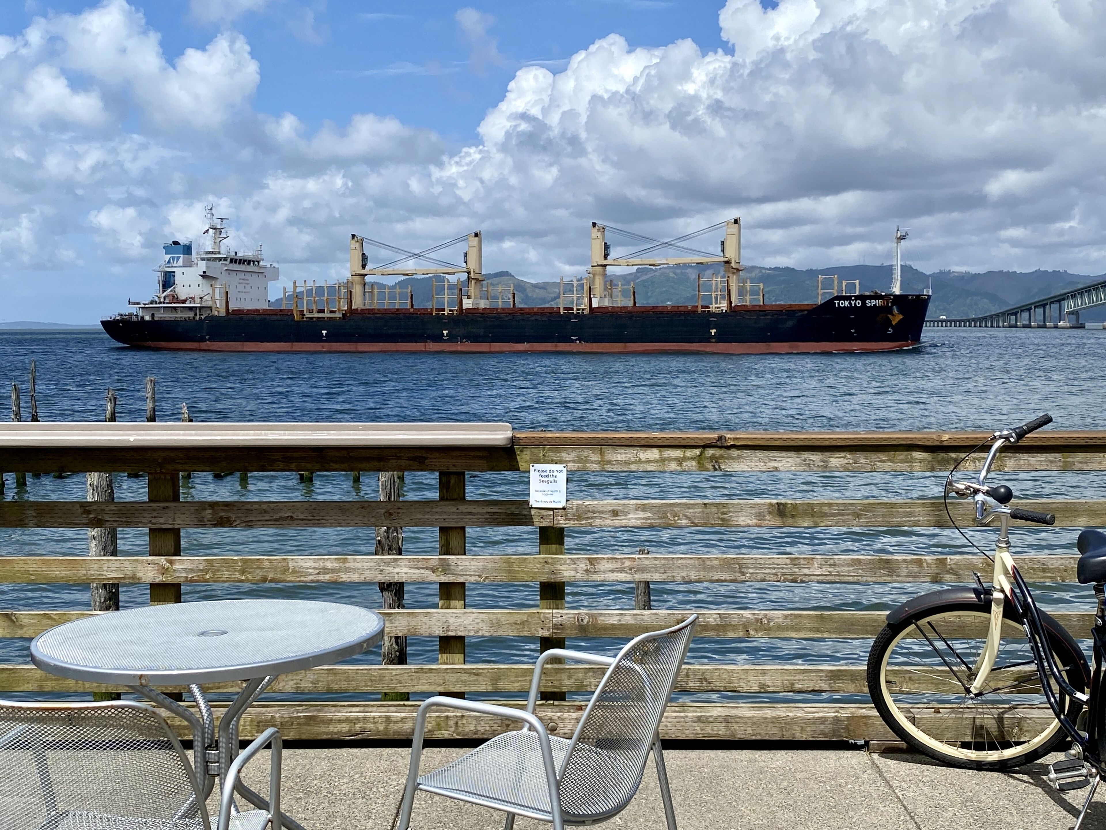 Une scène au bord de l’eau avec un grand cargo, un vélo et une table entourée de chaises.