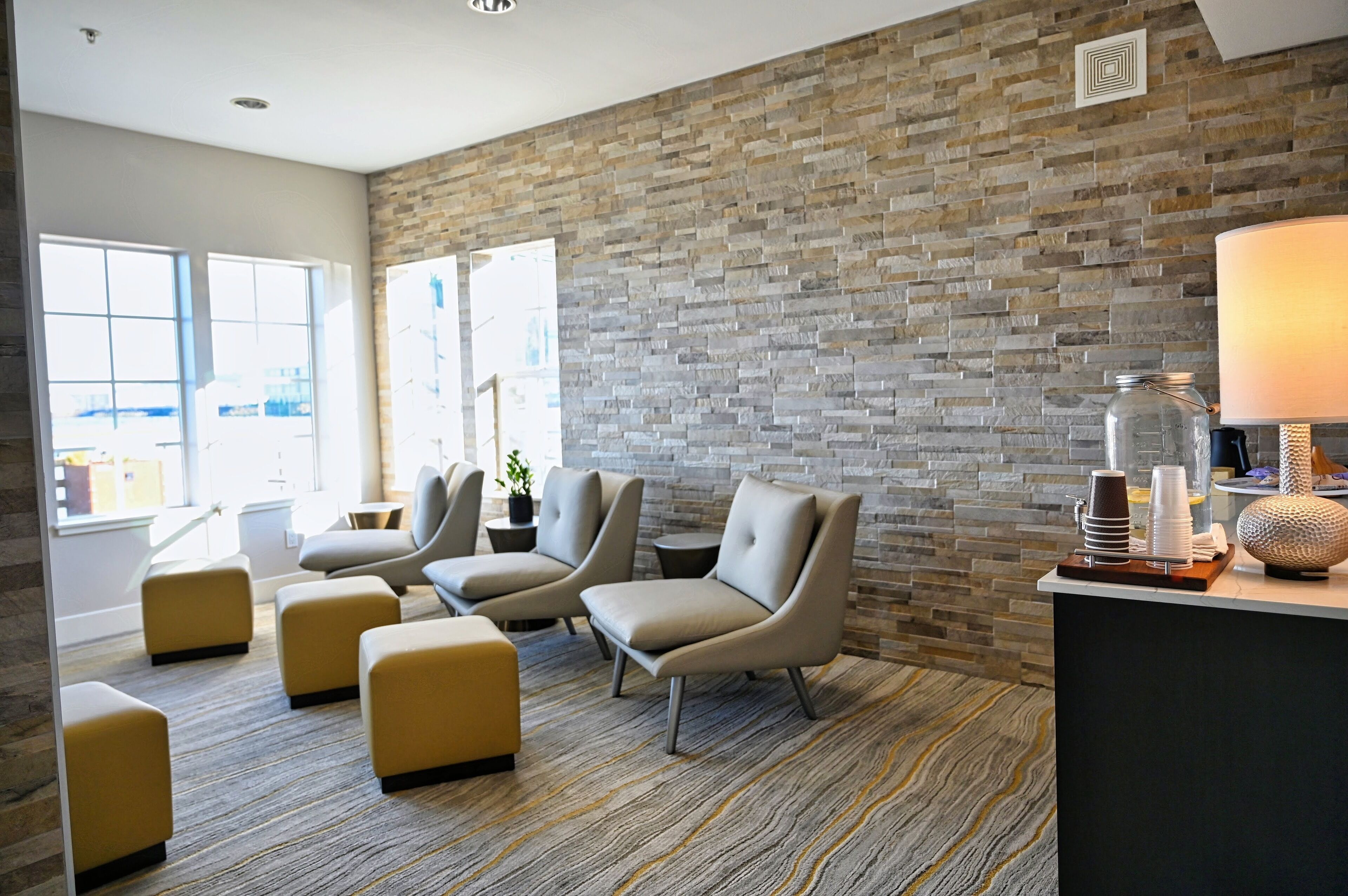 Un área de descanso moderna con sillones de cuero, alfombra estampada, pared de piedra y una recepción con lámpara y elementos decorativos.