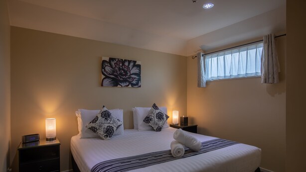 A neatly made bed with a striped bedspread, two pillows with floral patterns, a bedside lamp, and a wall-mounted artwork. - Abbella Lodge Motel (Christchurch)