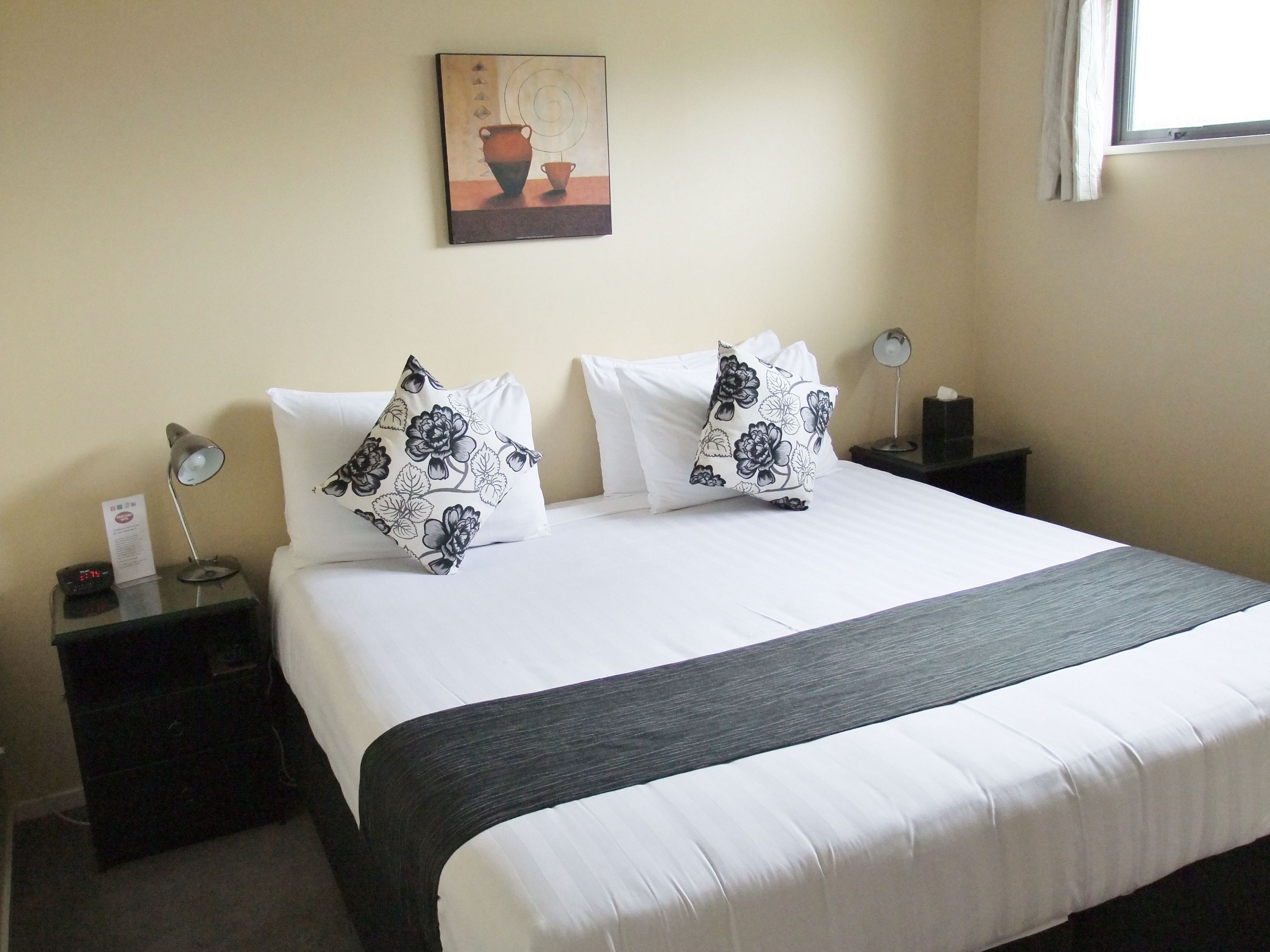 Una habitación de hotel con una cama grande, dos mesitas de noche, una mesilla auxiliar, un reloj de pared, un cuadro y una ventana con cortinas.