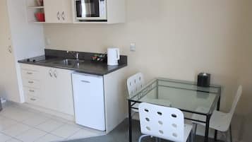 Cocina compacta con microondas, pileta y heladera pequeña. Hay una mesa de vidrio con dos sillas y una caja de pañuelos sobre la mesa.