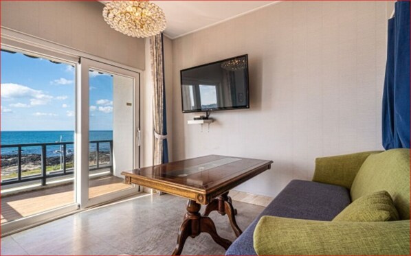 A room with a wooden table, a green sofa, a TV mounted on the wall, and a balcony with a view of the sea.