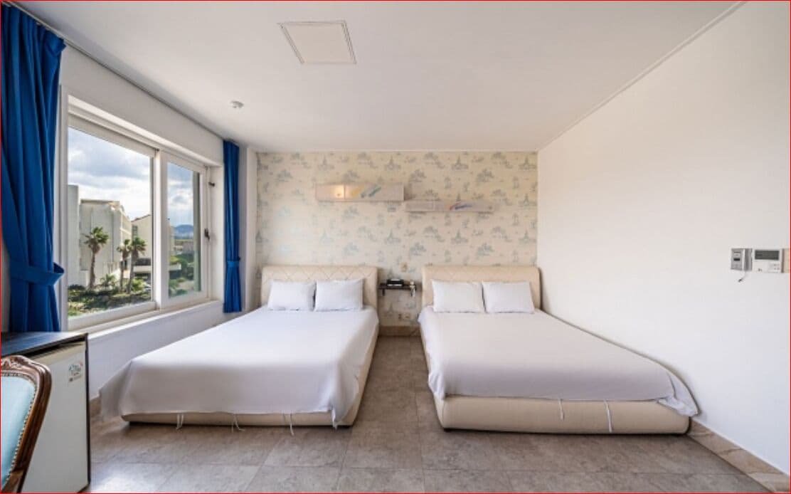A room with two single beds, a window with blue curtains, a wall-mounted air conditioning unit, and a view of palm trees and buildings outside.