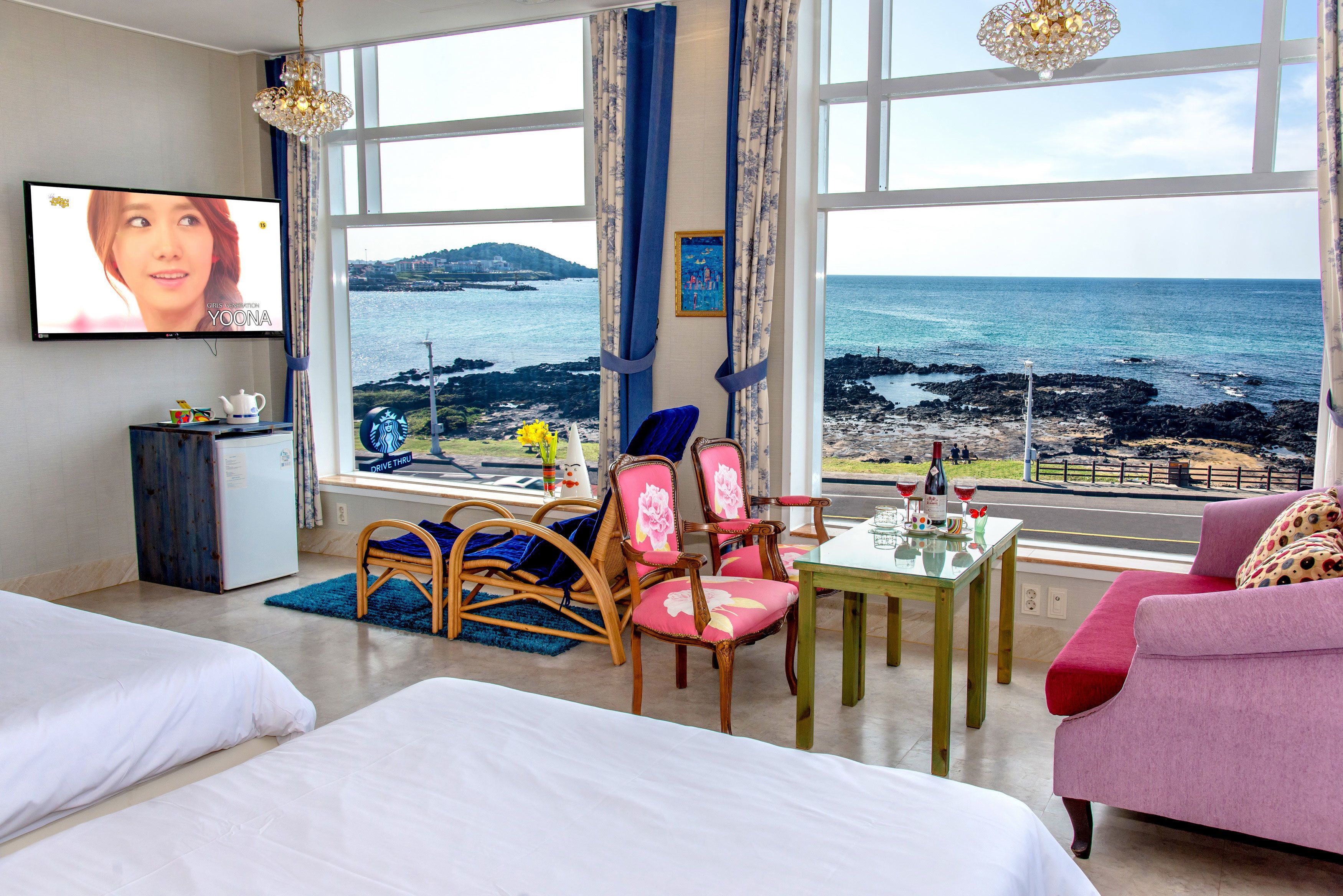 A hotel room with a large window offering a view of the sea and a distant island. There is a television mounted on the wall, a small table with a bottle of wine, and a pink sofa.