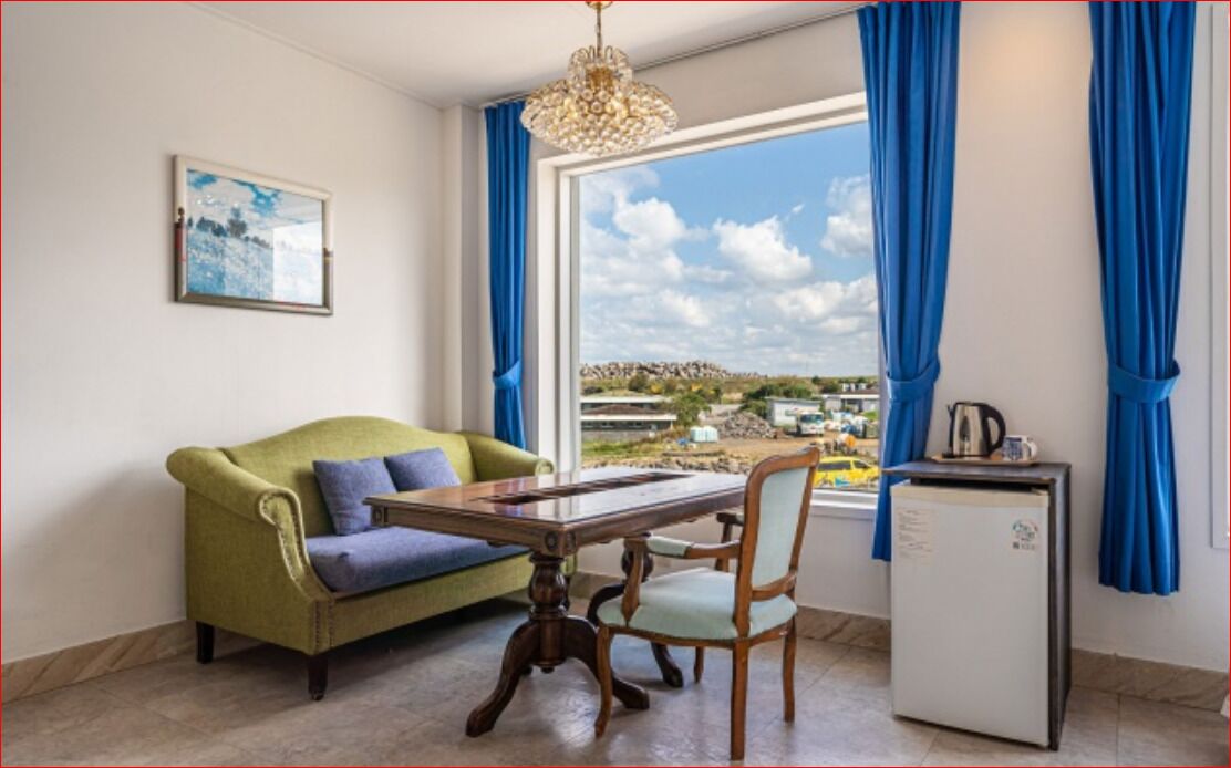 Ein Zimmer mit einem grünen Sofa, einem Holztisch, einem Stuhl, einer kleinen Kühlbox und einem Fenster mit blauen Vorhängen.