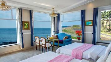 A hotel room with a large window offering a sea view, a bed, a chair, a small table with a bottle and fruit, and a vase with flowers.