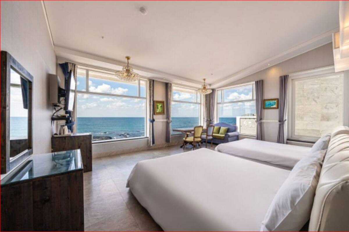 Habitació d'hotel amb un llit gran, una taula i una cadira, i vista al mar a través de grans finestres.