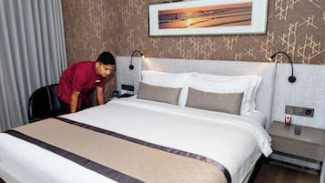 A hotel room with a neatly made bed, a painting above the headboard, and a nightstand with a phone and a red object.