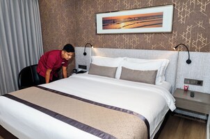 A hotel room with a neatly made bed, a painting above the headboard, and a nightstand with a phone and a red object.