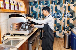 Ein Barista bereitet ein Getränk an einer Kaffeemaschine in einem Café mit strukturierter Wand und einer Neonreklame zu.