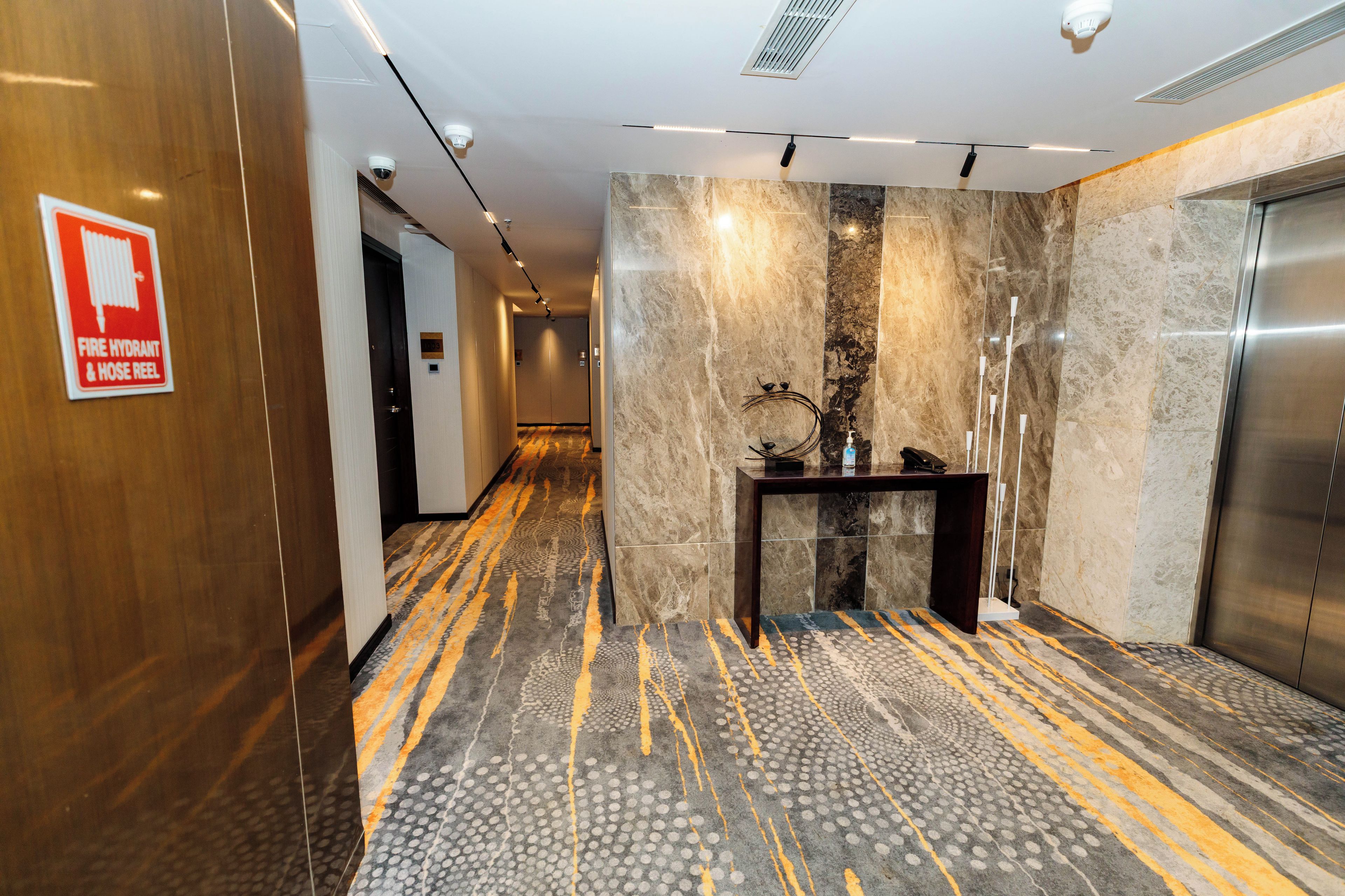 En hotelgang med brandhane og slangetromle-skilt, en marmorvæg, et mønstret tæppe og en elevator.