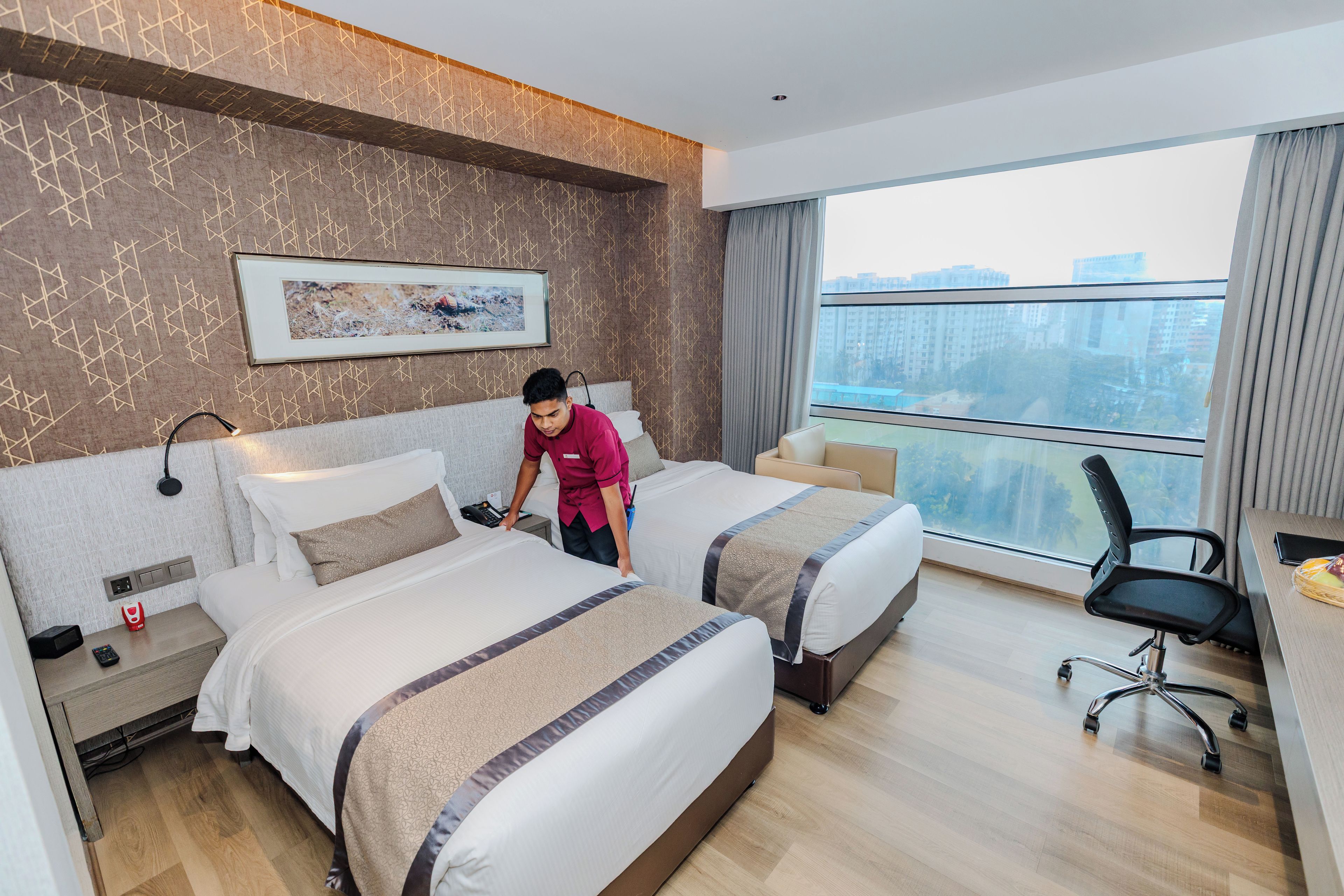 A hotel room with two beds, a desk, a chair, and a large window with a city view.