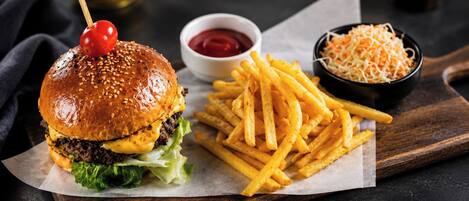 A burger with a sesame seed bun, a beef patty, cheese, lettuce, and a cherry tomato on a skewer, accompanied by French fries and two dipping sauces.