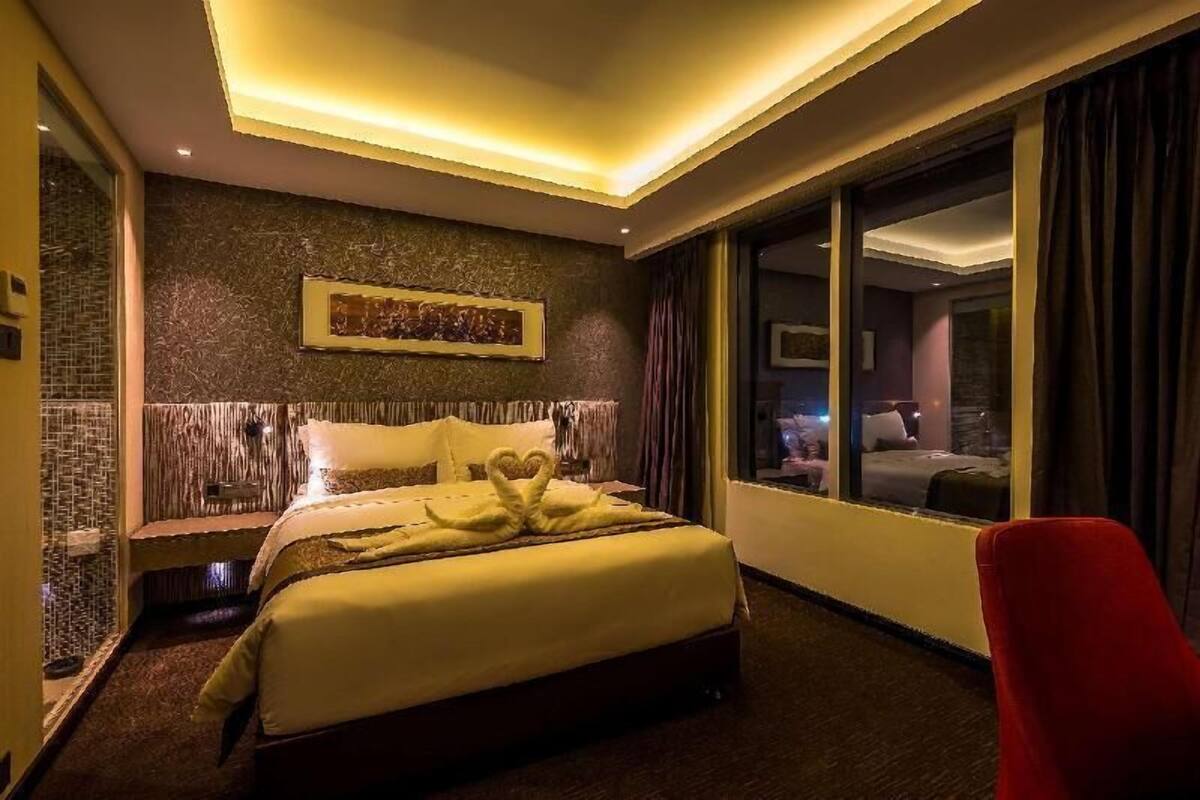 Quarto de hotel com uma cama, uma cadeira vermelha, uma janela grande e uma obra de arte na parede.