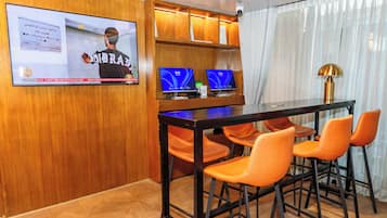 Een vergaderzaal met een houten paneelwand, een groot scherm waarop een nieuwsuitzending draait, een bureau met twee monitoren en oranje stoelen.