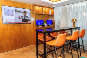 Un salón de conferencias con pared de paneles de madera, una pantalla grande mostrando un noticiero, un escritorio con dos monitores y sillas naranjas.