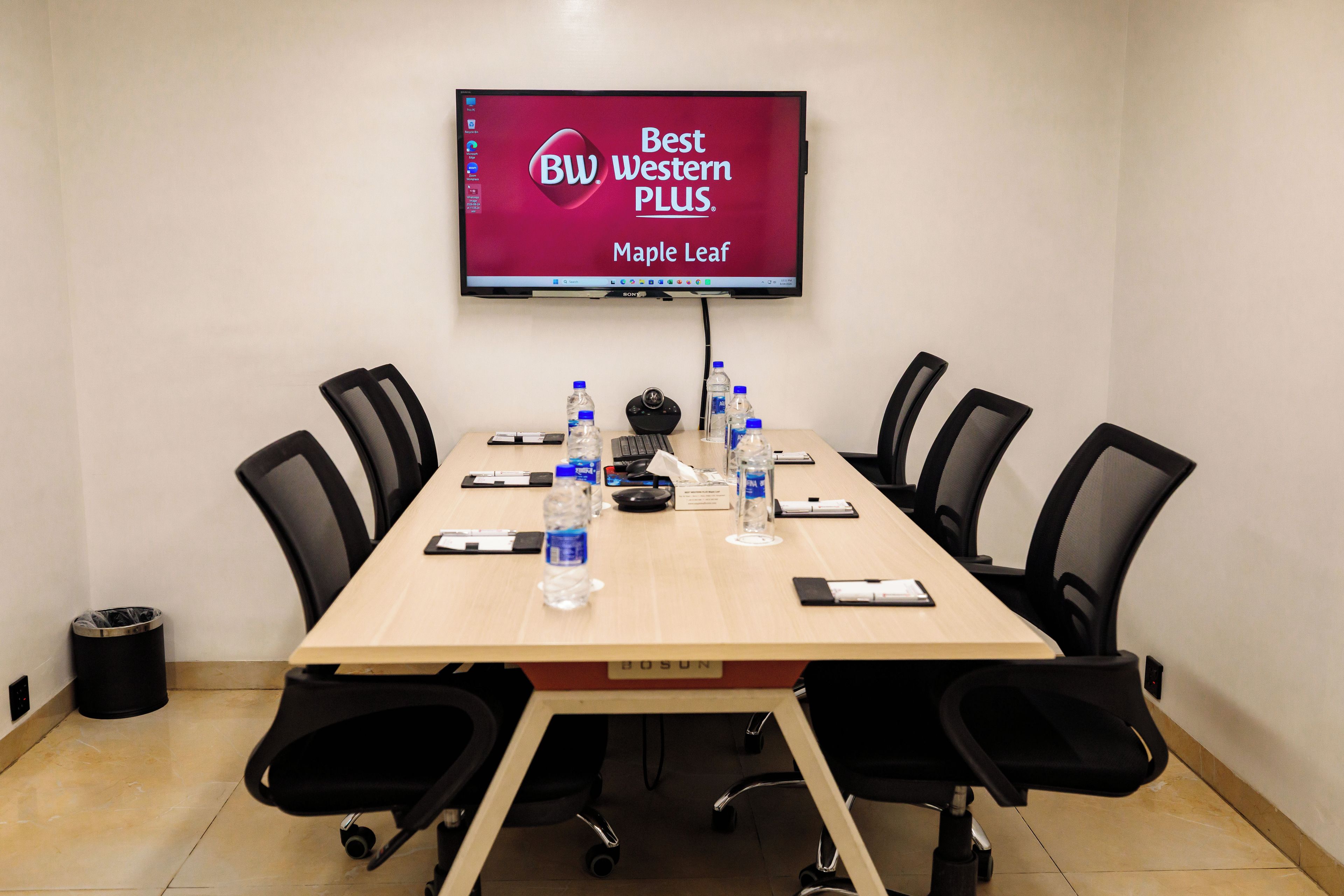A conference room with a long table, black chairs, a mounted TV displaying 'Best Western PLUS Maple Leaf', and water bottles on the table.