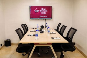 Ein Konferenzraum mit einem langen Tisch, schwarzen Stühlen, einem an der Wand montierten Fernseher, der 'Best Western PLUS Maple Leaf' anzeigt, und Wasserflaschen auf dem Tisch.