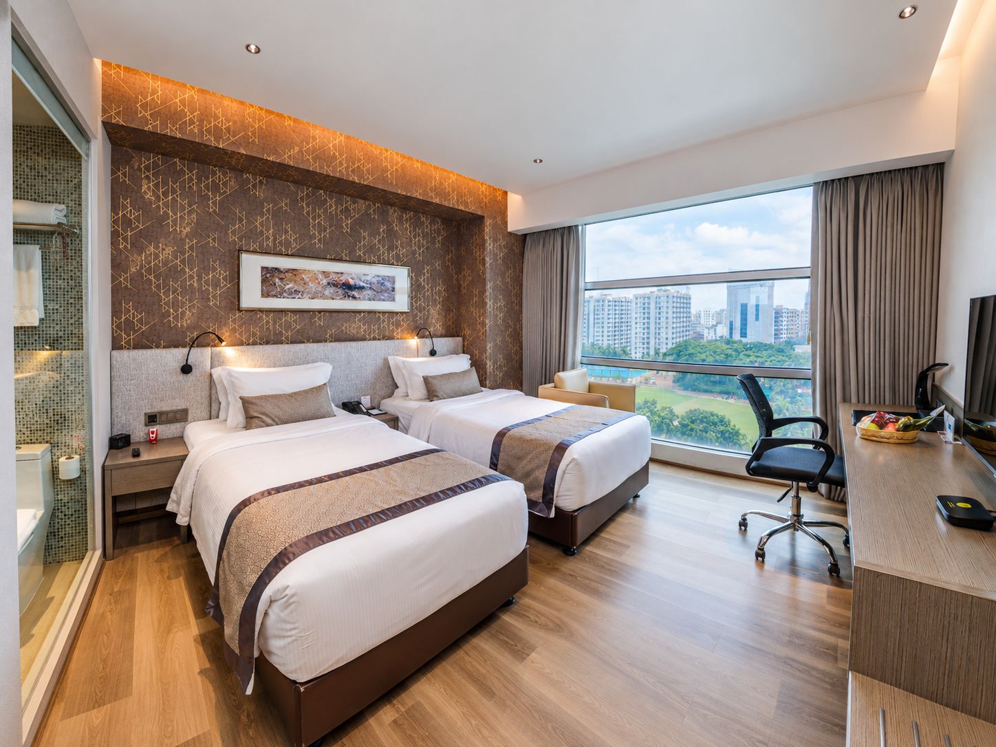 A hotel room with two beds, a desk, and a large window offering a city view.