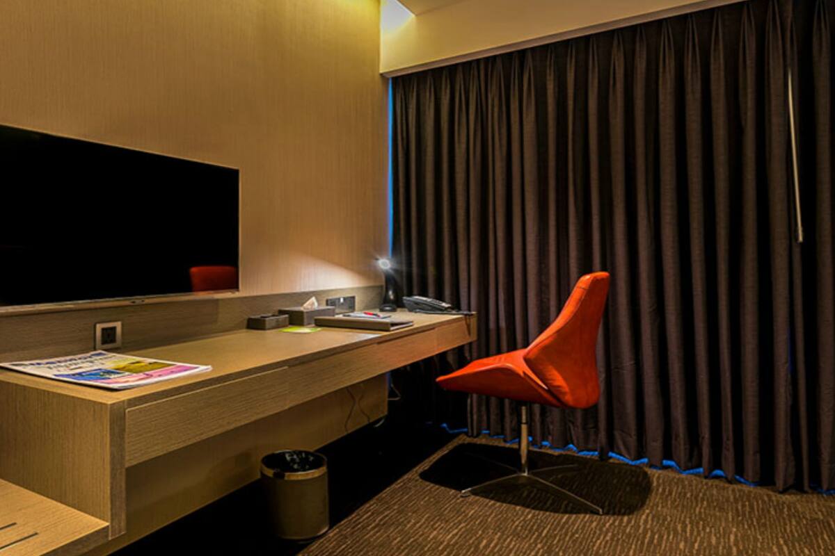 Quarto de hotel com escrivaninha de madeira, cadeira vermelha, televisão de tela plana e uma janela grande com cortinas escuras.