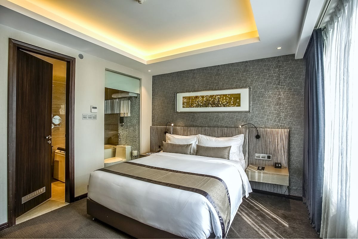 Quarto de hotel moderno com uma cama grande, uma mesa de cabeceira com abajur, cabeceira com alto-falantes embutidos e uma obra de arte pendurada na parede.