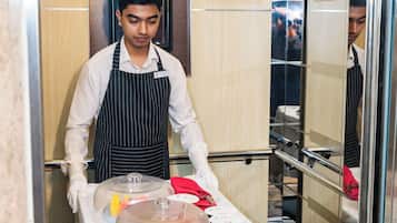 A hotel staff member in a white shirt and black apron, preparing a table with food items and condiments.