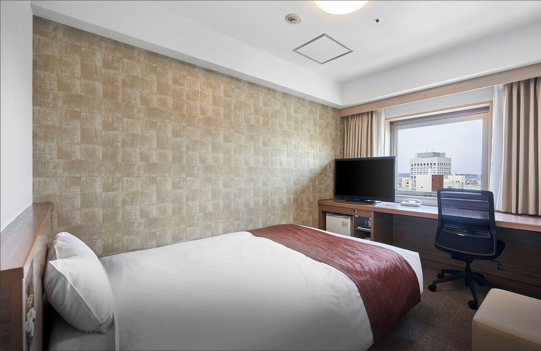 A hotel room with a bed, a desk with a television, a chair, and a window with a city view.