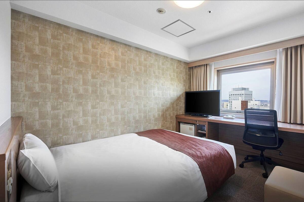 A hotel room with a bed, a desk with a television, a chair, and a window with a city view.