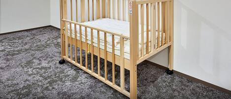 A wooden crib with a mattress, wheels, and a safety guard, placed on a carpeted floor in a room with white walls.