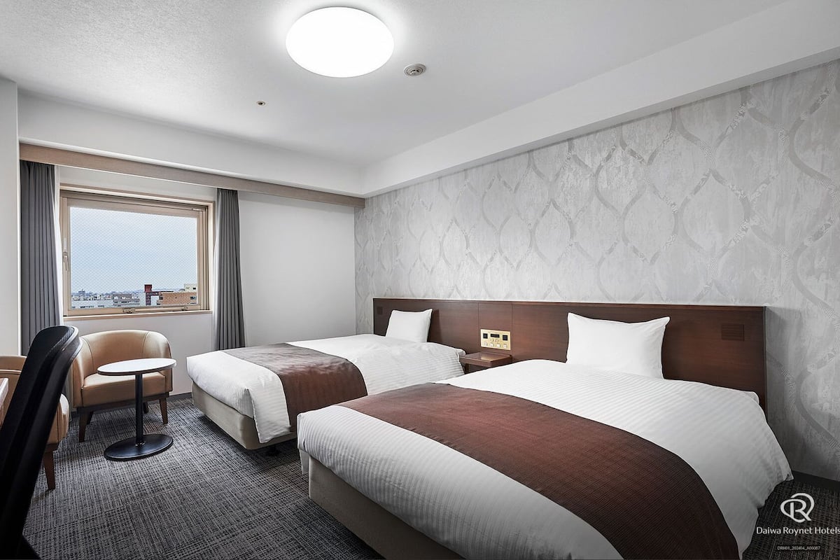 A hotel room with two beds, a desk, a chair, a TV, and a window with a city view.