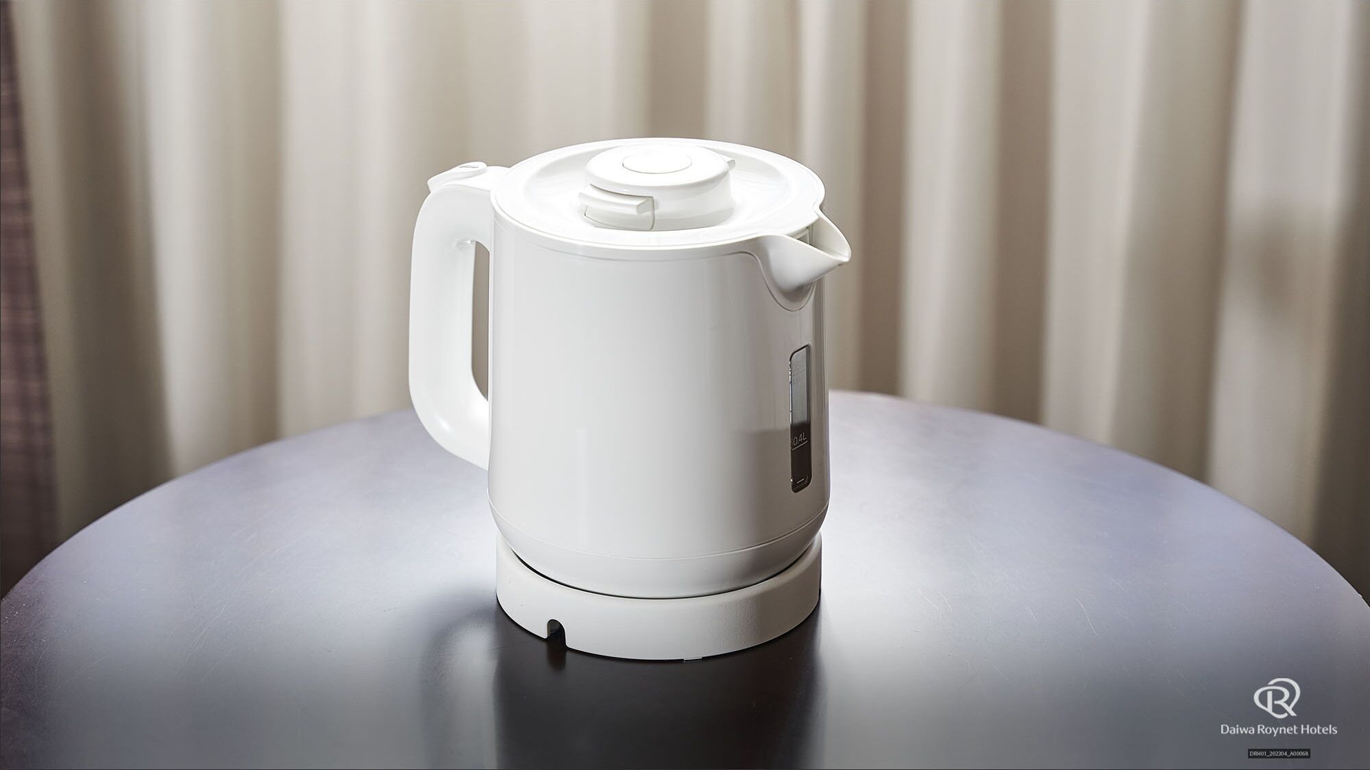 Una tetera eléctrica blanca sobre una mesa redonda con el logotipo de Daiwa Roynet Hotels.