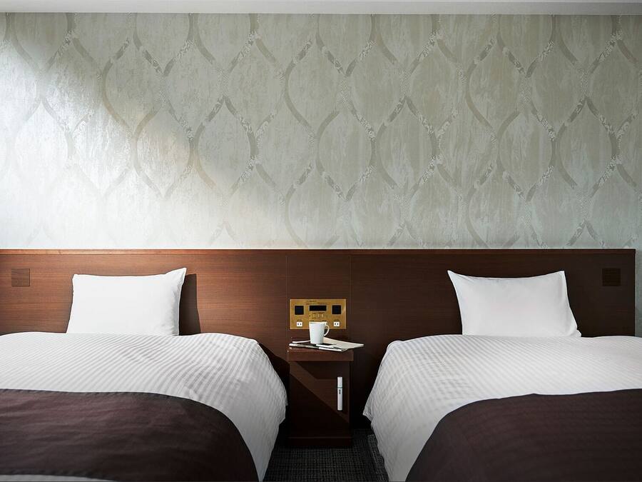 A hotel room with two beds, a nightstand with a lamp, and a wall with a patterned wallpaper.