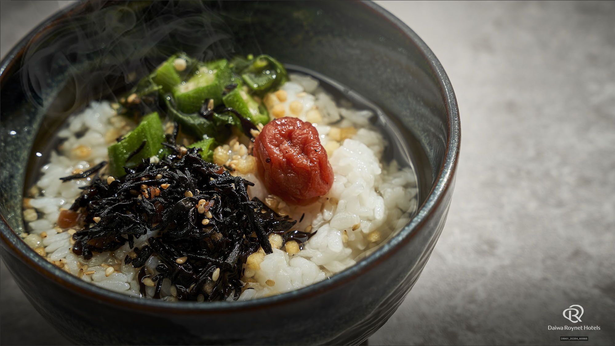 A bowl of rice with seaweed, green vegetables, and a red fruit, possibly a tomato.