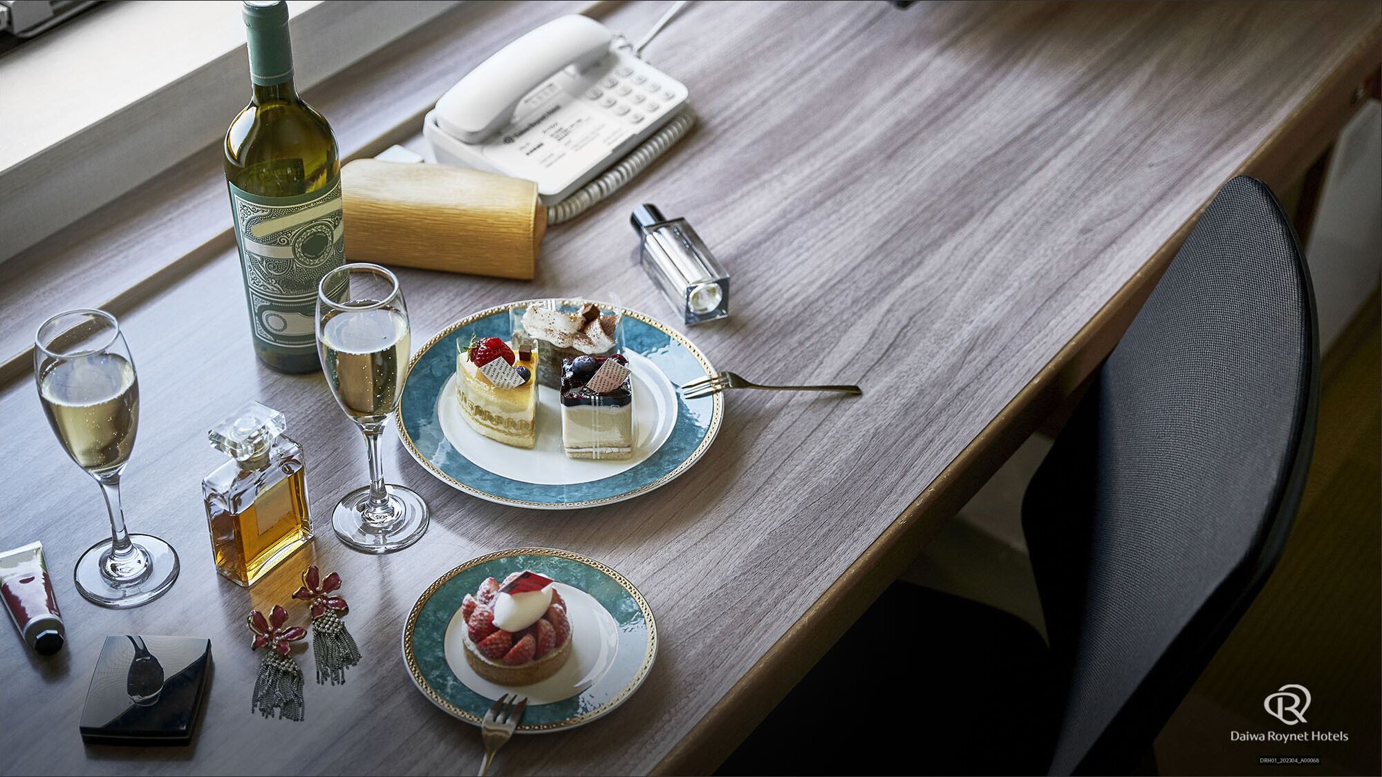 A table set with two plates of desserts, a bottle of wine, glasses, a telephone, and a pen.