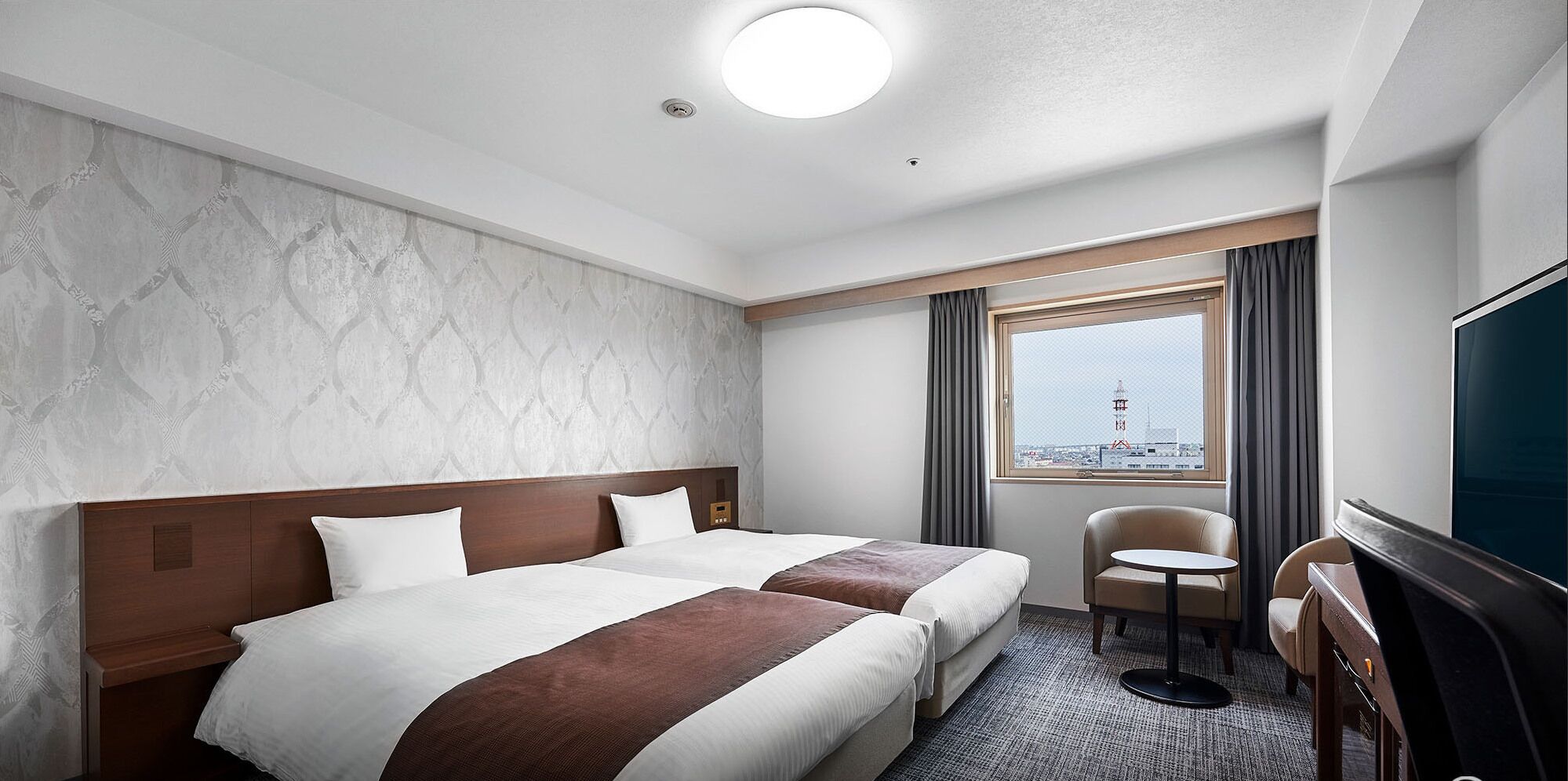 A hotel room with two beds, a desk, a chair, a TV, a window with a view, and a ceiling light.