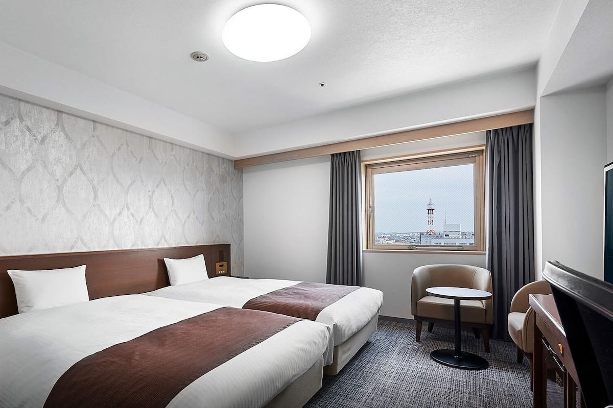 A hotel room with two beds, a desk, a chair, a TV, a window with a view, and a ceiling light.