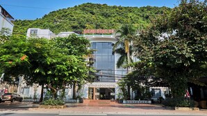 A hotel with a modern facade, a crosswalk in front, and a lush green hill in the background. - Gieng Ngoc Hotel (Hai Phong)