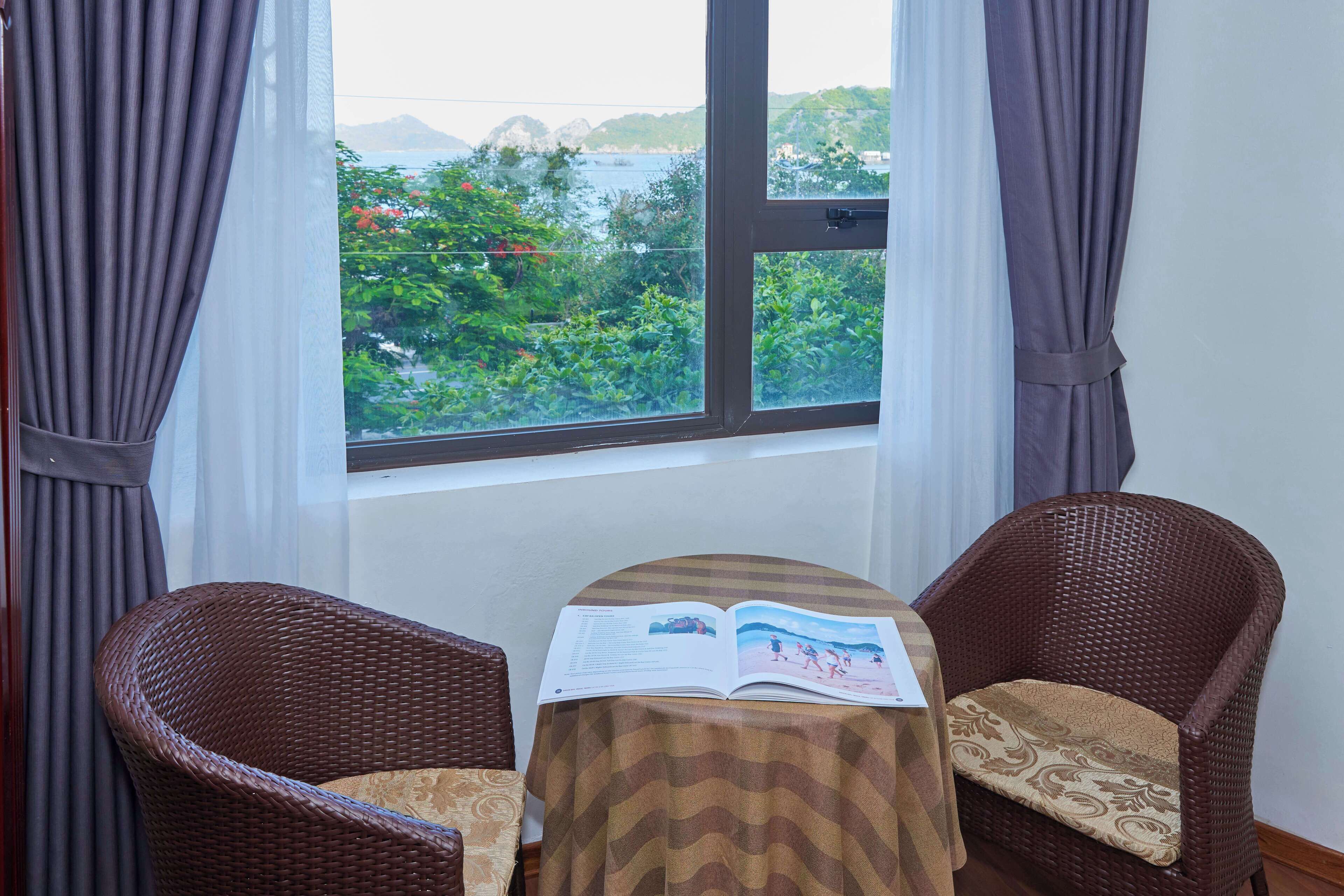 A room with a large window, two wicker chairs, a table with a book, and a view of greenery and mountains.