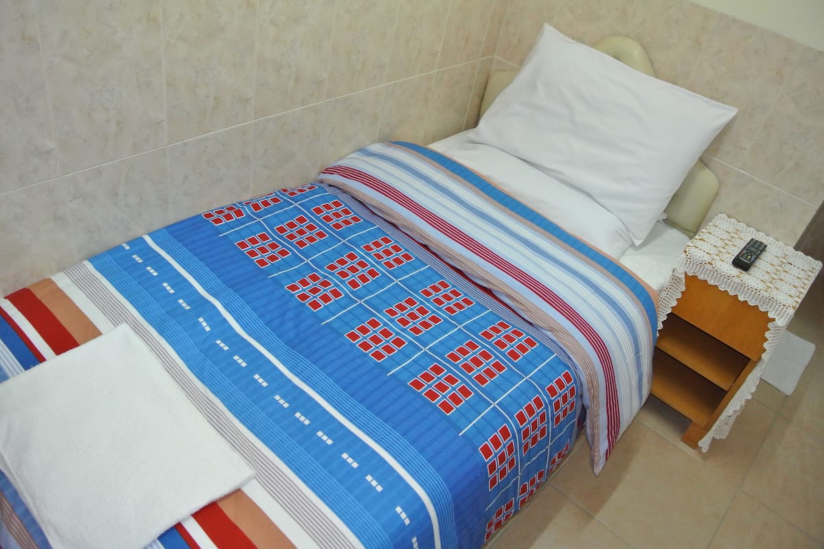 Un lit bien fait, recouvert d’une couvre-lit à motifs bleus et rouges, avec un oreiller blanc et une table de chevet en bois sur laquelle se trouve une télécommande.