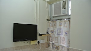 A bathroom with a mounted television, an air conditioning unit, a shower curtain with a floral pattern, and tiled walls. - Fortunate Guest House (Kowloon)