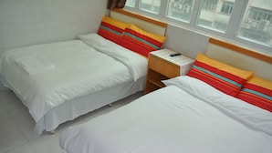 A hotel room with two beds, each with white bedding and colorful striped pillows. There is a wooden nightstand with a remote control and a small table between the beds. - Fortunate Guest House (Kowloon)